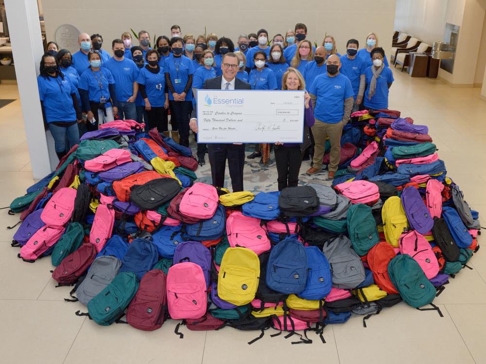 A group of people in blue shirts stands behind a large pile of colorful backpacks, holding a $50,000 donation check from Essential Foundation.