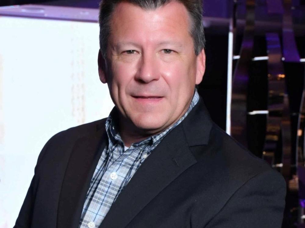 Headshot of a man in a dark blazer and checkered shirt, smiling slightly, with a blurred modern interior background.