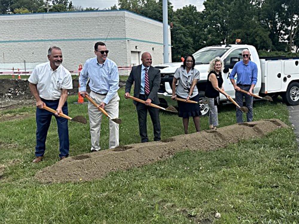 Six people with shovels participate in a groundbreaking ceremony on a grassy area, with a building and a white utility truck in the background.