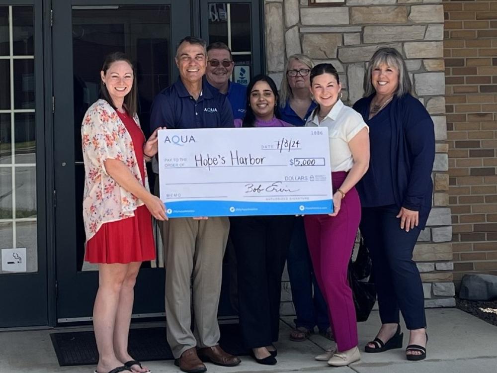 Group of people holding a large check for $5,000 made out to Hope’s Harbor, presented by Aqua, standing in front of a building.