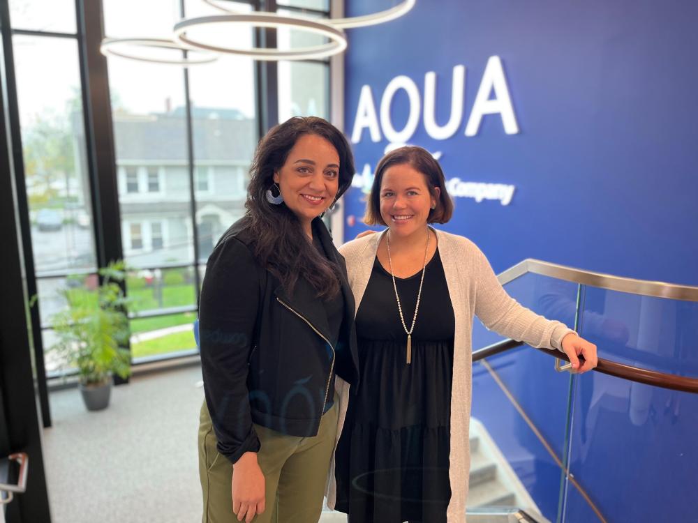 Two women standing on a staircase in front of a blue wall with the AQUA logo, smiling and posing together inside an office building.