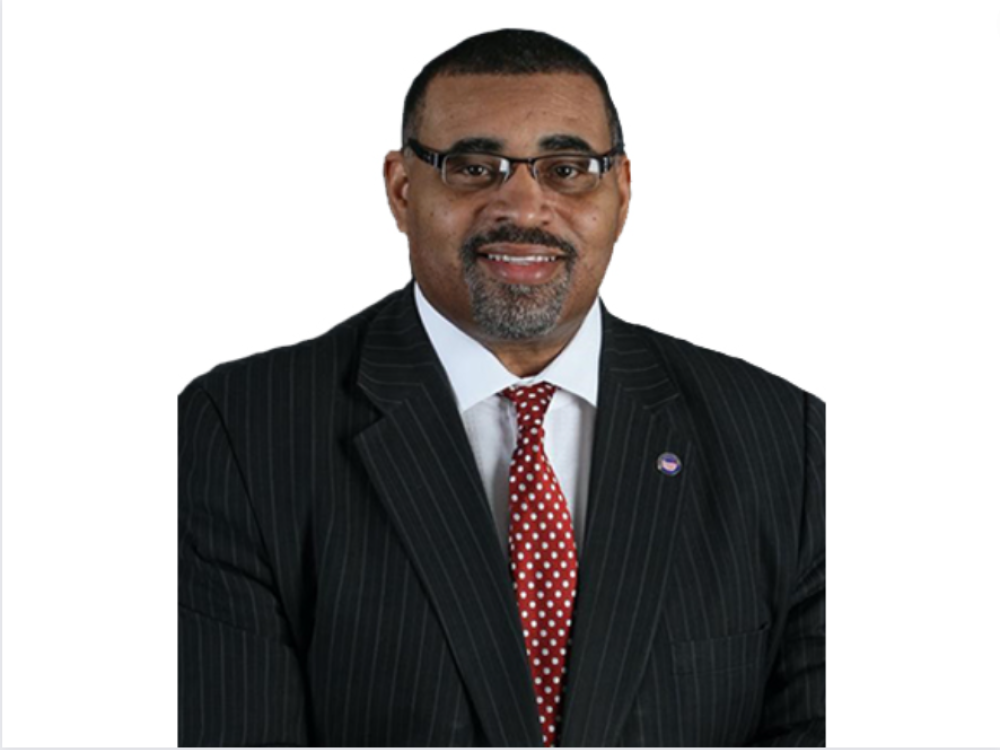 Man wearing glasses, a pinstripe suit, and a red polka-dot tie, smiling against a plain white background.