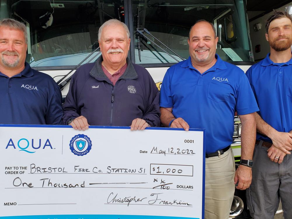 Four men, including Aqua representatives, hold a large $1,000 donation check for Bristol Fire Company, standing in front of a fire truck.