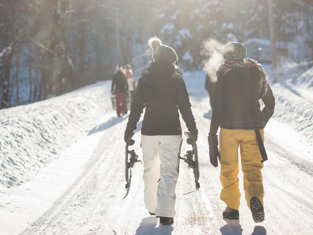 Two people in winter gear walk down a snowy path carrying snowshoes, with their breath visible in the cold air and sunlight.