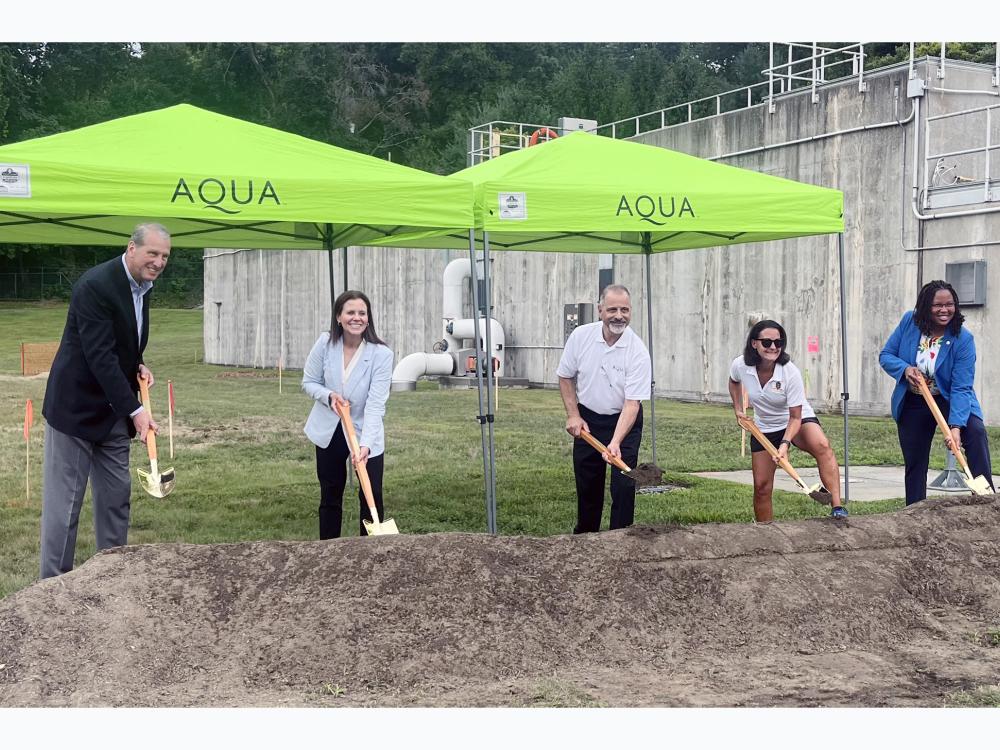 Five people with shovels participate in a groundbreaking ceremony under two green tents with the Aqua logo, near a concrete structure.