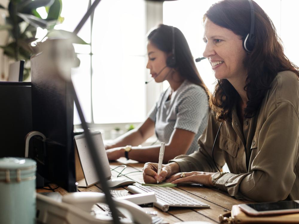 Two women wearing headsets work at computers in an office, with one smiling and taking notes while the other is focused on her screen.