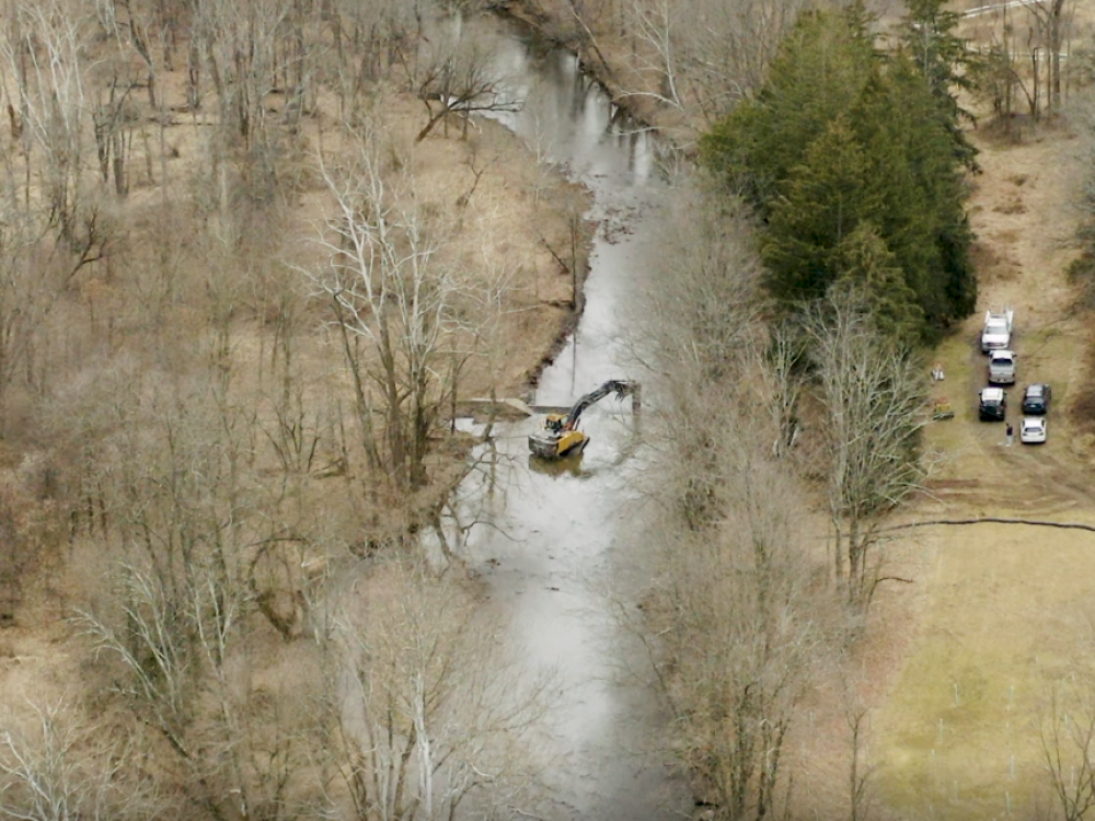 An aerial view of a construction vehicle working in a stream surrounded by bare trees, with trucks parked nearby on a grassy area.