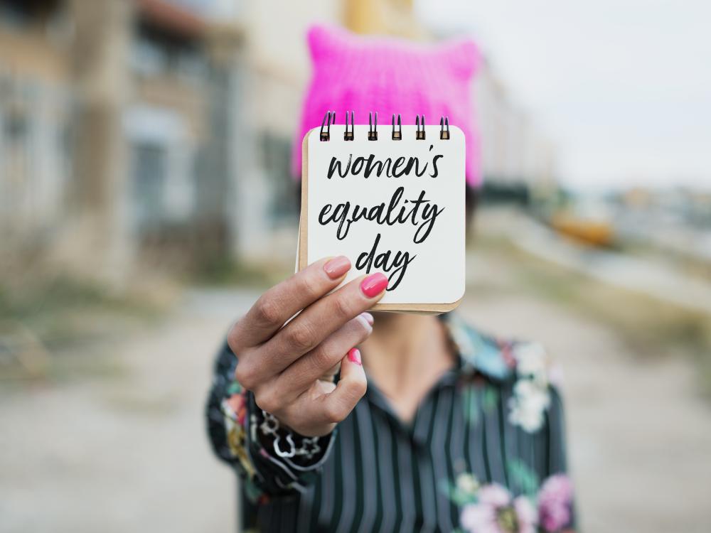 Person wearing a pink hat holds a notepad with 'women's equality day' written on it, with a blurred urban background.