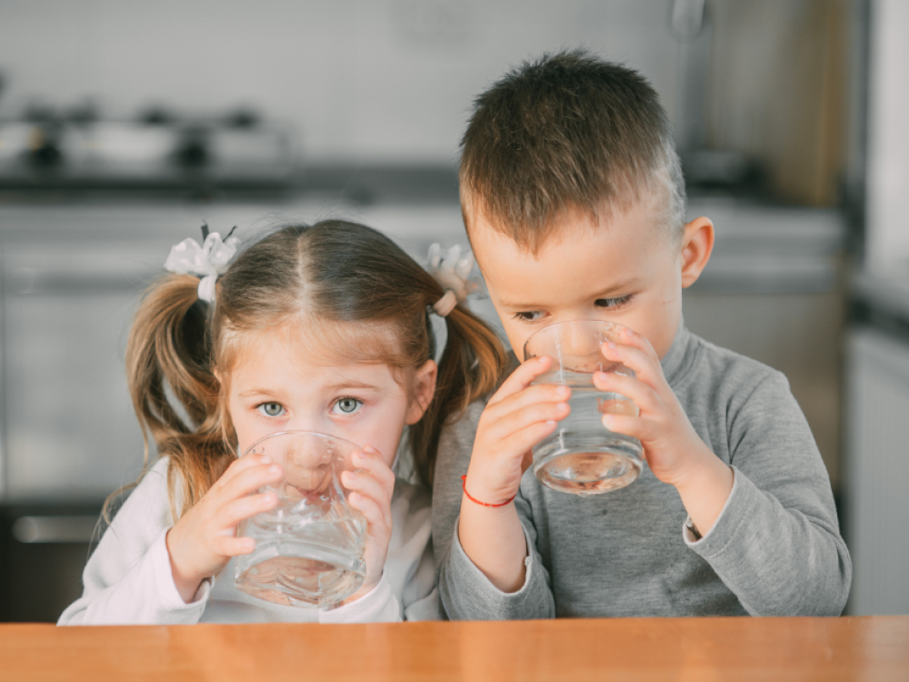  Two young children sitting at a table, drinking water from clear glasses, with a blurred kitchen background.