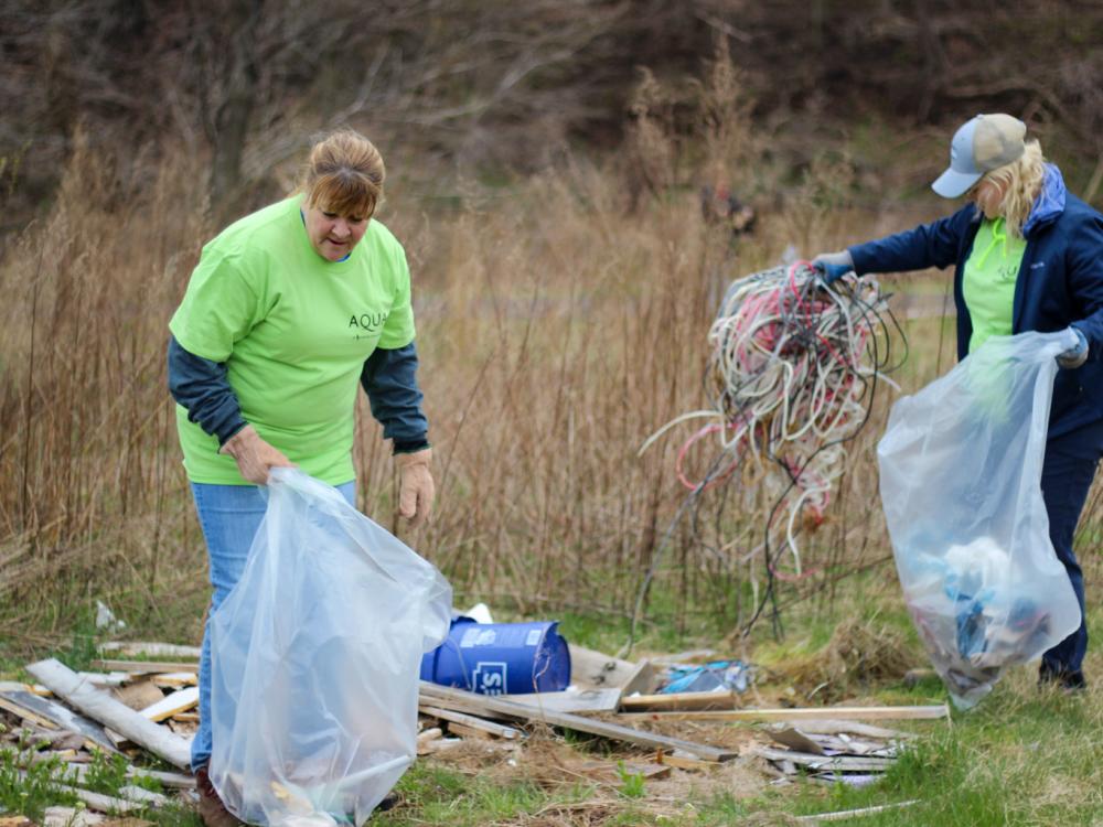 Two volunteers in green shirts pick up debris and wires during an outdoor cleanup event, collecting trash in large plastic bags.