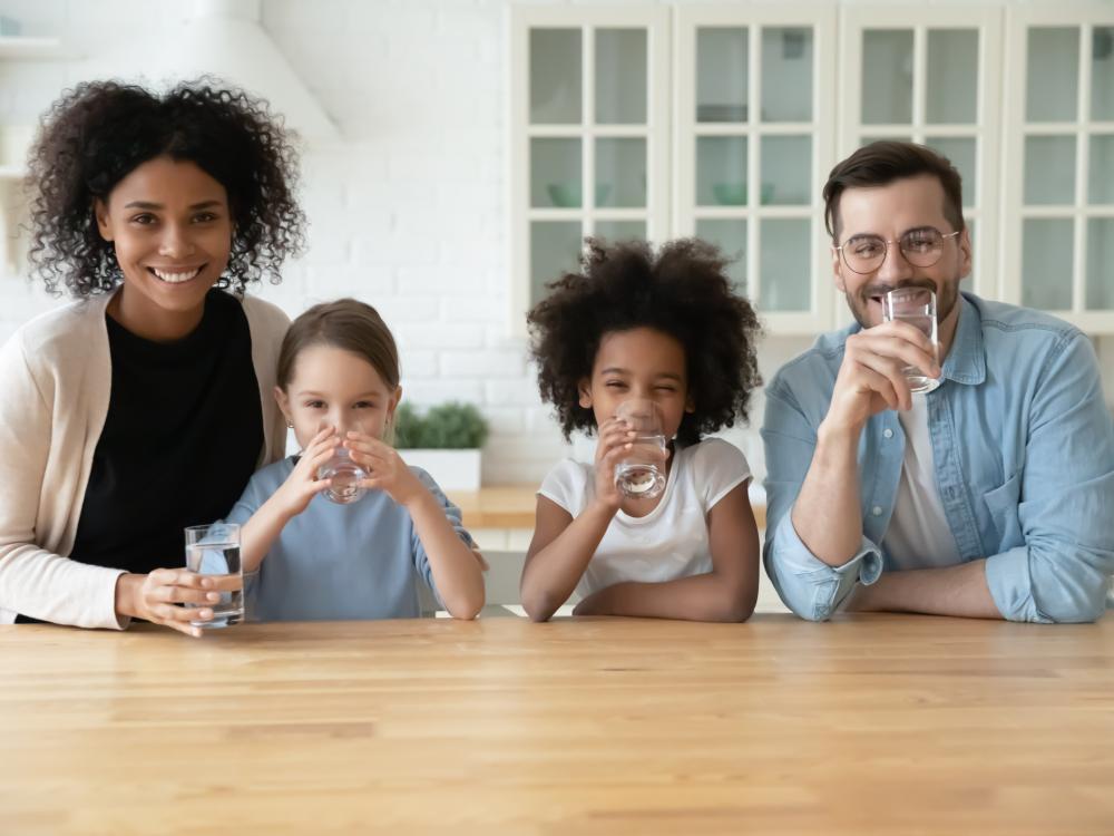 A smiling family of four sits at a kitchen table, each drinking a glass of water, creating a cheerful and healthy atmosphere.