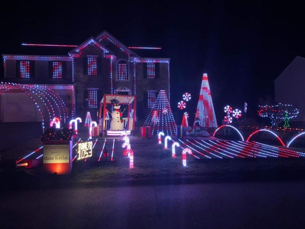 A house decorated with elaborate Christmas lights, featuring a snowman, candy canes, and a sign promoting donations to Make-A-Wish.