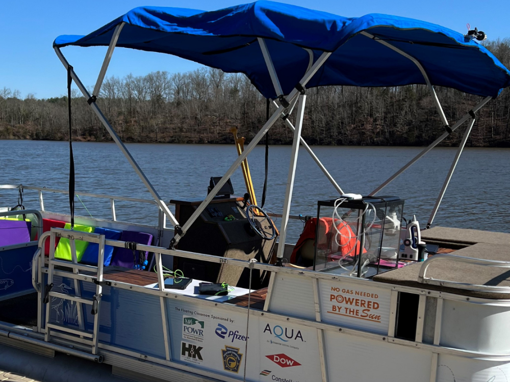 Pontoon boat on a lake with seats and a canopy, labeled 'Powered by the Sun,' sponsored by various companies including AQUA.