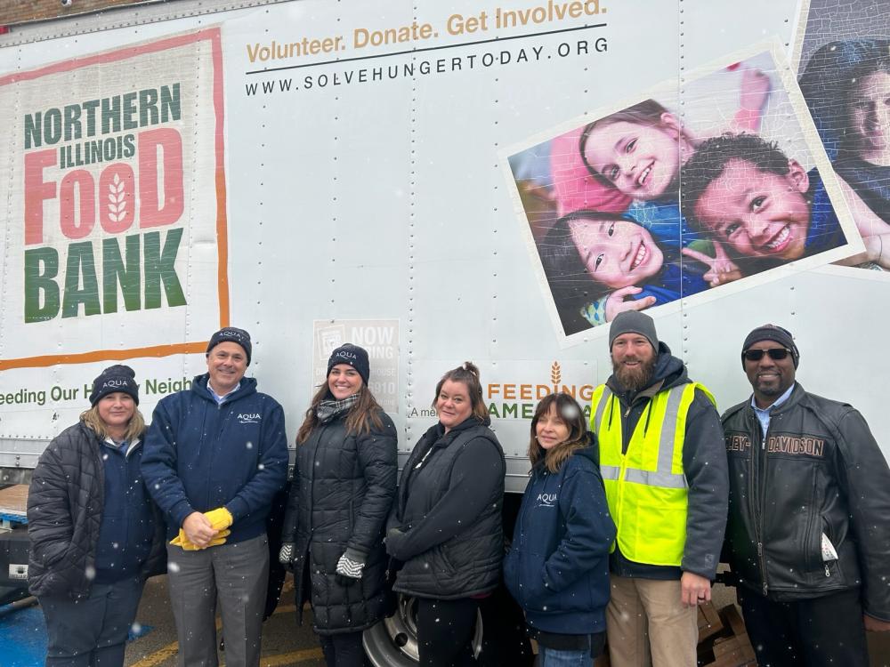 A group of volunteers from Aqua stand in front of a Northern Illinois Food Bank truck, smiling together on a snowy day.