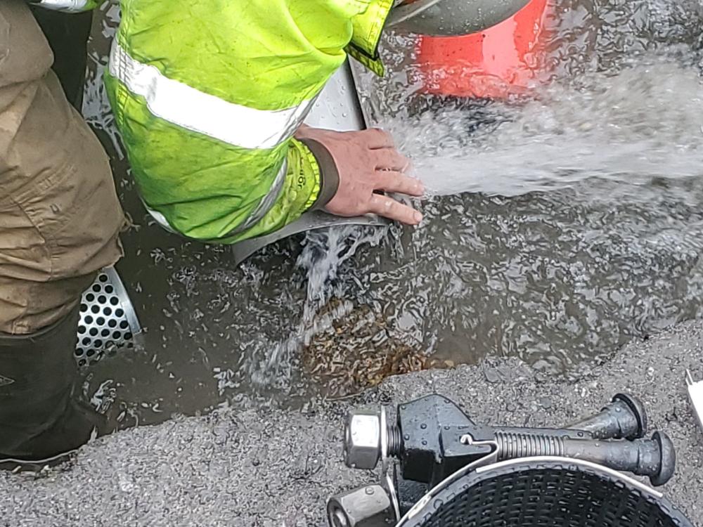 Close-up of a worker in a reflective jacket handling water flowing from a pipe, standing in a puddle, performing maintenance work.