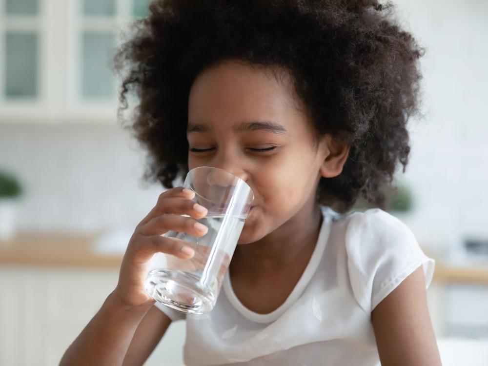 A young child with curly hair drinks a glass of water, smiling with eyes closed, in a bright kitchen setting.