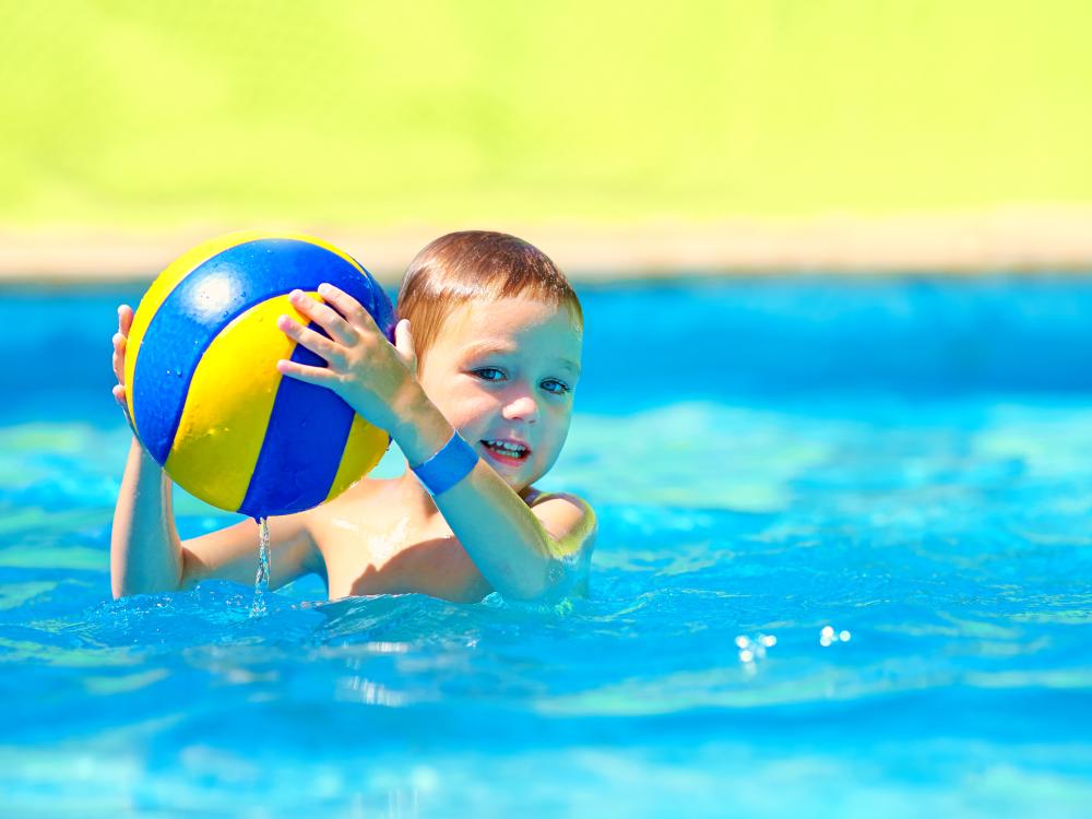 A young child plays in a pool, holding a yellow and blue ball, with a bright green background in the distance.