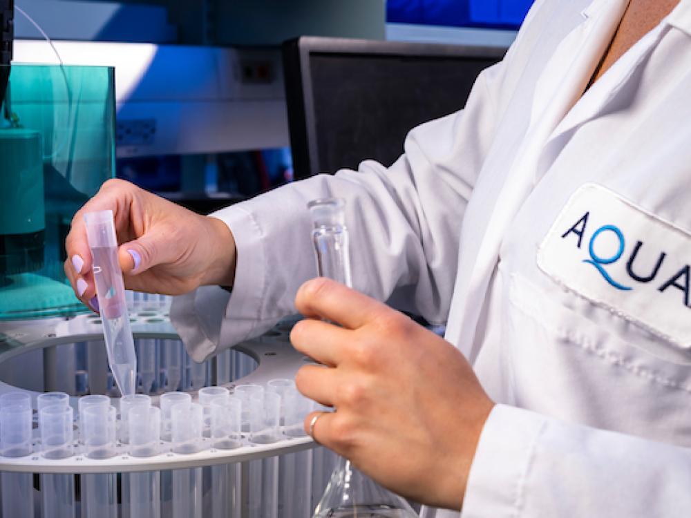 A lab technician in a white coat labeled "AQUA" handles test tubes and a flask in a laboratory setting with specialized equipment.