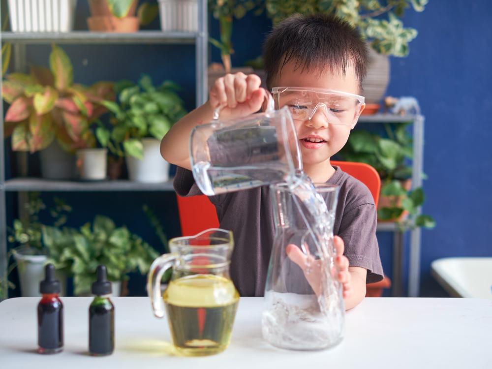 A young child wearing safety glasses pours water into a glass jar while conducting a science experiment at home with colorful liquids.