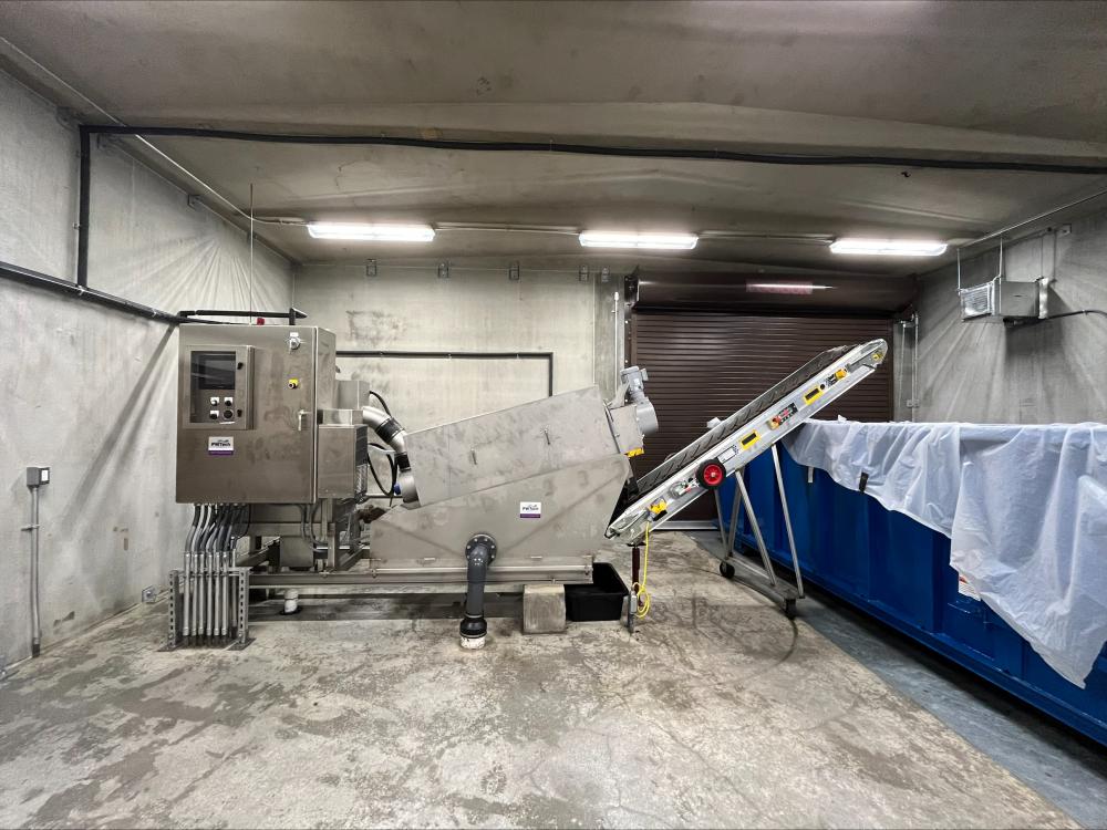 Industrial equipment in a concrete room with a conveyor belt leading to a large blue container covered in plastic.