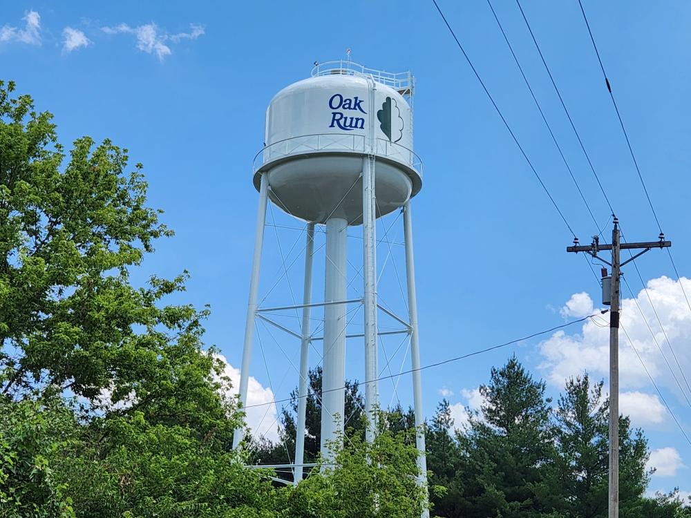 Water tower labeled 'Oak Run' with a tree logo, surrounded by green trees and power lines, set against a clear blue sky.