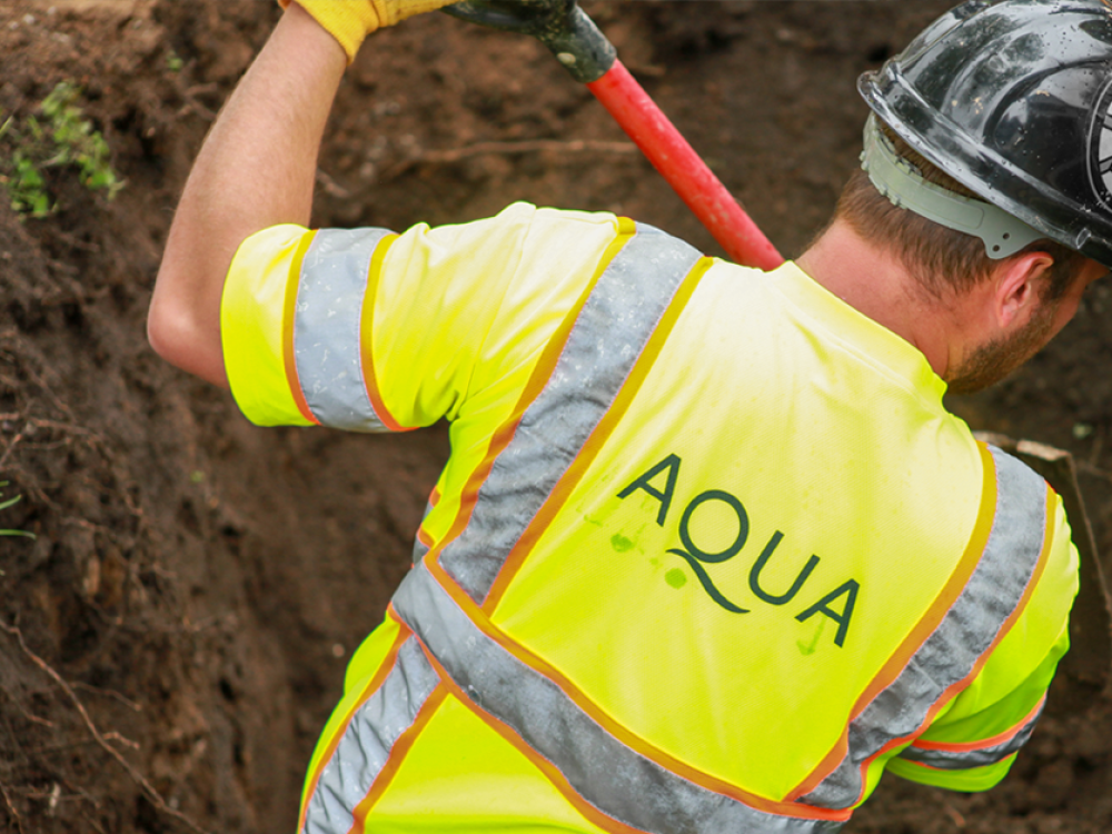 Construction worker in a high-visibility vest and hard hat, with 'AQUA' logo on the back, working with a shovel in a trench.