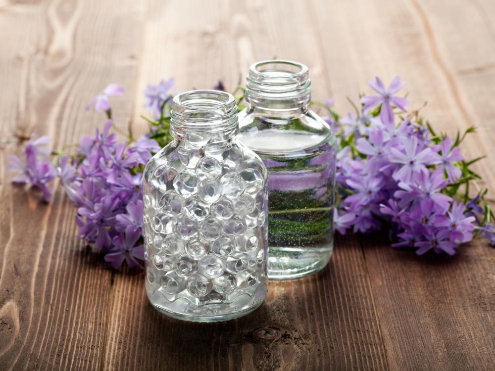 Two glass bottles, one filled with clear beads and the other with liquid, surrounded by purple flowers on a wooden surface.