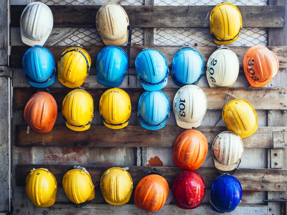 Various colored hard hats, including yellow, blue, white, orange, and red, hanging on a wooden board, representing safety gear.