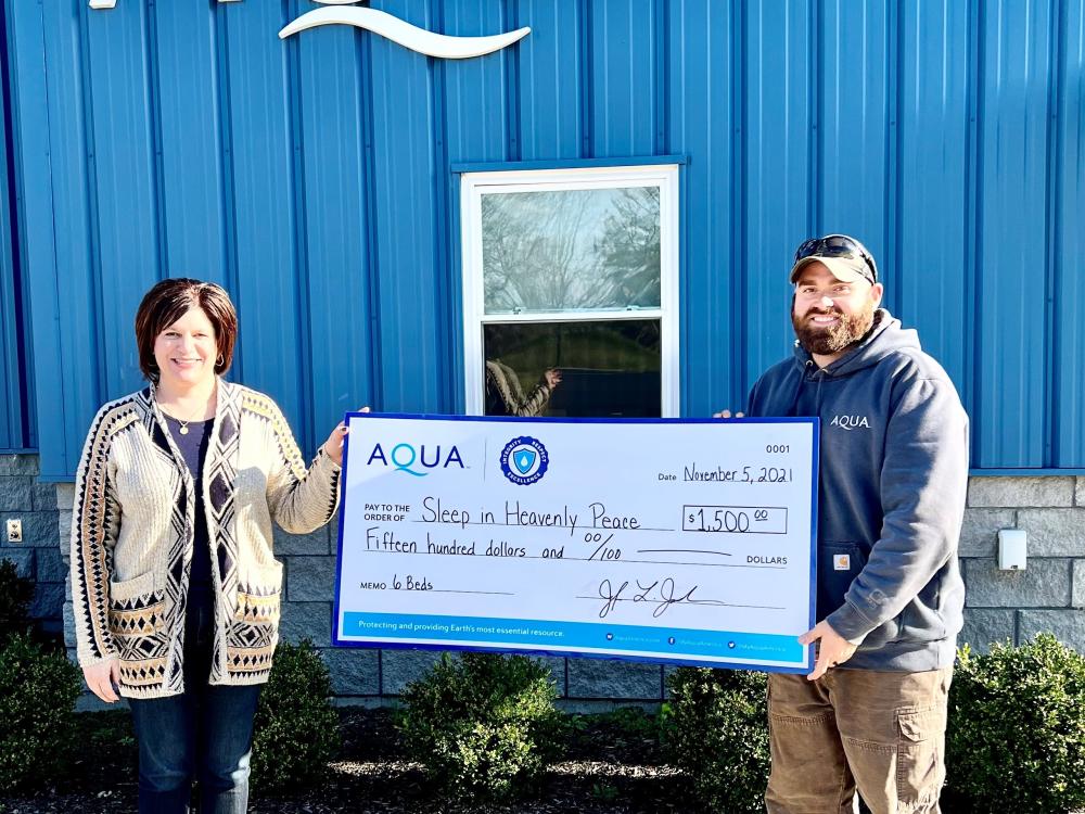 Two people holding a large check for $1,500 in front of an AQUA building, supporting the organization Sleep in Heavenly Peace.