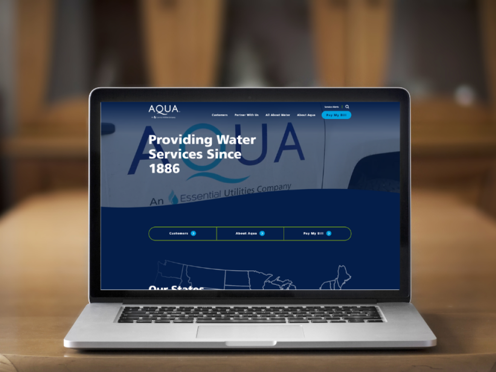 Laptop displaying the AQUA website with the text 'Providing Water Services Since 1886' on the screen, set on a wooden table.