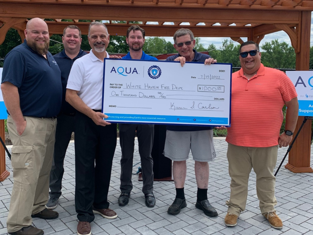 Six men holding a large check for $1,000 from AQUA, addressed to White Haven Fire Dept., standing outside under a wooden structure.