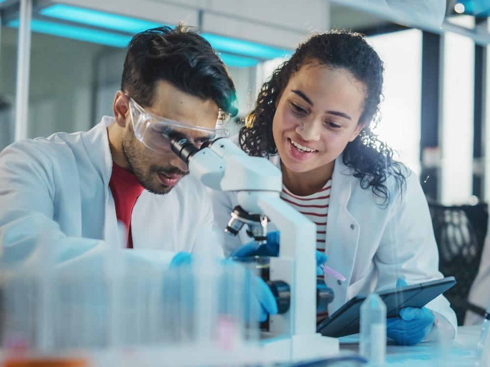 Two scientists wearing lab coats and goggles work together in a laboratory, observing a sample through a microscope.