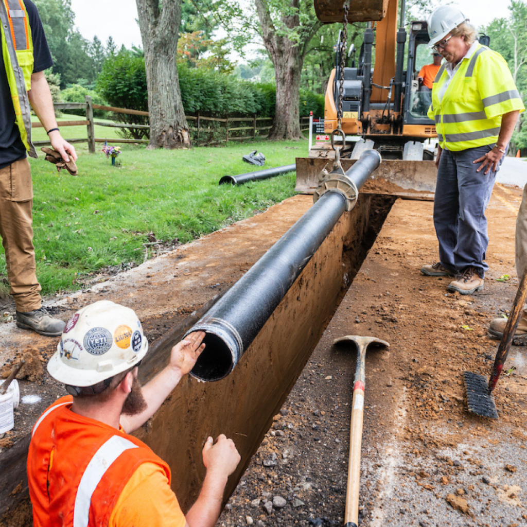 Three construction workers are installing a large black pipe in a trench on the side of a road. 