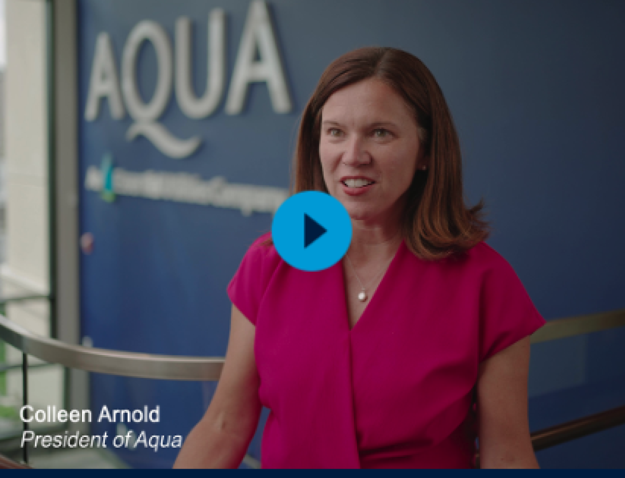 Brown haired woman in fuchsia blouse, standing in the lobby of a building with an Aqua sign on the wall behind her