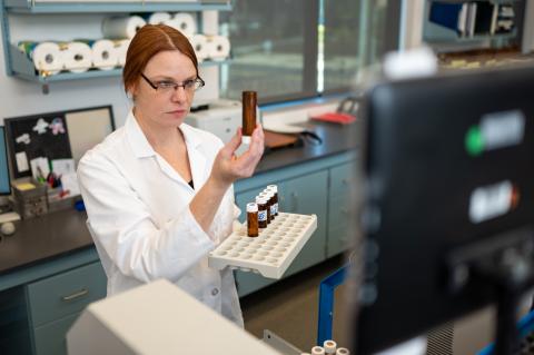 Women with red hair in laboratory, holding a test tube and tray in front of a computer, front view