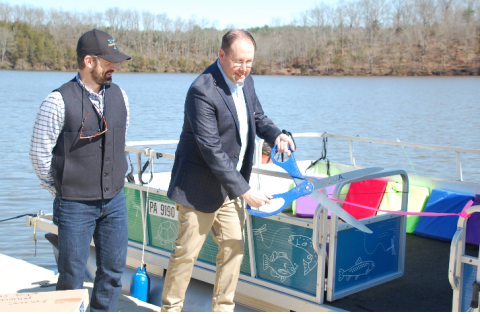 Two people next to a pontoon boat on a river, cutting a ribbon with a large scissors. 