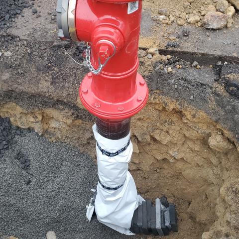 Close-up of a red fire hydrant being installed in a trench, with the base wrapped and secured, surrounded by gravel and soil.