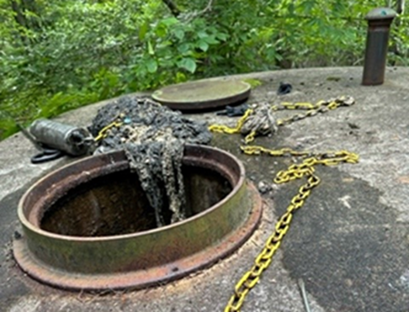 Open tank with sludge spilling out, yellow chains, and dense foliage in an outdoor setting.