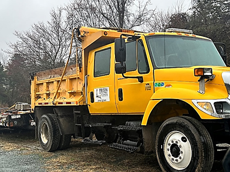 Yellow dump truck parked on gravel, with trees in the background and a contractor sign on the door.