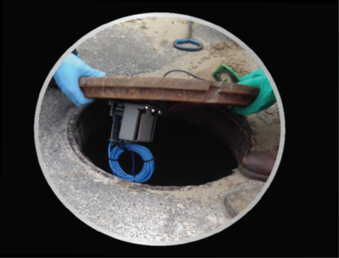 A worker wearing gloves lifts a manhole cover equipped with a sensor device and blue wiring underneath.