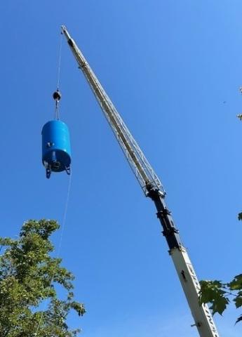 A crane lifts a blue tank high into the air against a clear blue sky, with tree branches visible in the foreground.