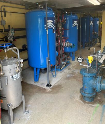 Interior view of a water treatment facility with multiple blue filtration tanks, pipes, and a 'Raw Water Bag Filter' unit in the foreground.